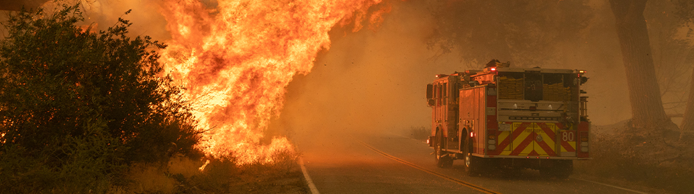 A fire truck passes a large brush fire on the side of the road.