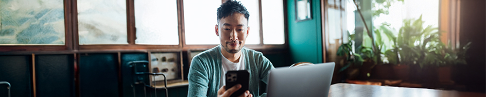Man looks at his phone while at his desk with his laptop open.